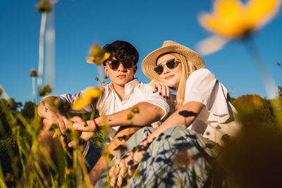 From below cheerful woman and boyfriend in sunglasses smiling and looking at camera while spending time on sunny day in nature