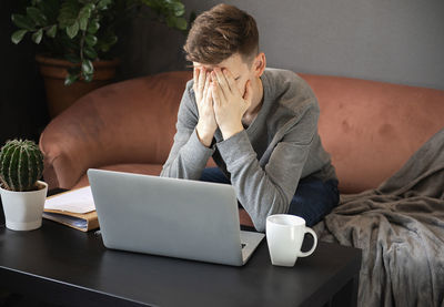 Frustrated young man student looking exhausted and covering his face with hands  sitting at laptop 