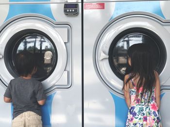 Rear view of siblings standing against washing machines at laundromat