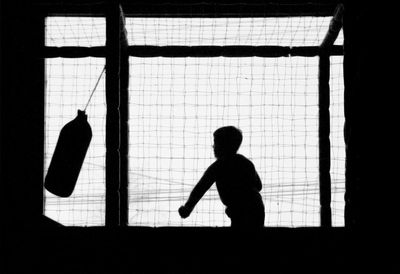 Silhouette boy punching on bag hanging by window