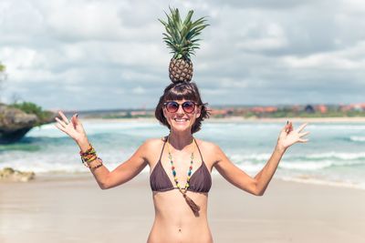 Portrait of happy young woman in bikini balancing pineapple on head at beach