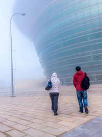 Full length rear view of man and woman walking against building during foggy weather