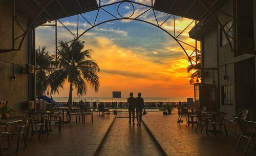 People on sidewalk by sea against sky during sunset