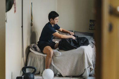 Teenage boy with backpack sitting on bed in bedroom at home