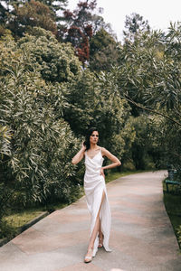 An elegant young woman bride in a wedding dress walks through a green park among plants and trees