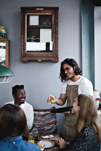 Smiling owner serving food to young man and women at dining table