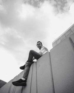 Low angle view of young man sitting on wall against sky