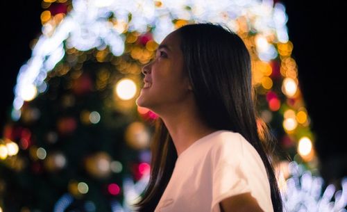 Young woman looking up while standing against illuminated christmas tree at night
