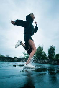 Full length of young woman jumping in puddle against sky