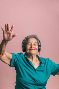 Joyful modern senior female listening to music in headphones and dancing with closed eyes on pink background in studio