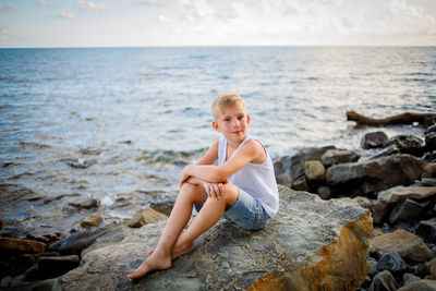 Boy sitting on rock by sea against sky