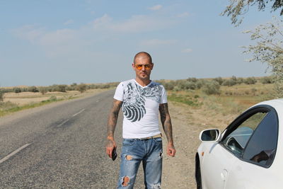 Man with tattoo standing by car on street against sky