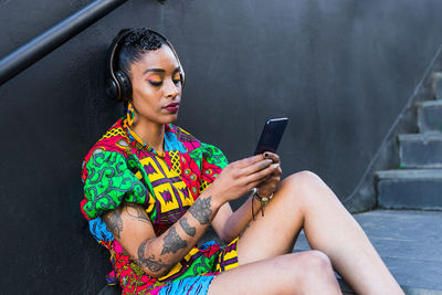 Young ethnic female in colorful stylish outfit listening to music in wireless headphones and browsing social media on cellphone