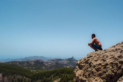 Shirtless man crouching on cliff against clear sky