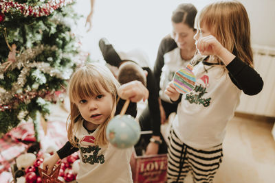 Girl showing christmas ornament at camera while family decorating christmas tree in home