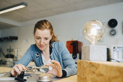 Confident redhead female engineer looking through magnifying glass at workbench in creative office