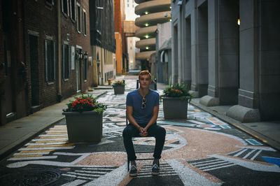 Portrait of young man sitting on chair at footpath amidst buildings