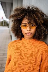 Beautiful ethnic woman with curly afro hair wearing vivid orange sweater looking at camera against gray wall