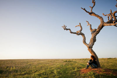 Woman sitting by bare tree against clear sky