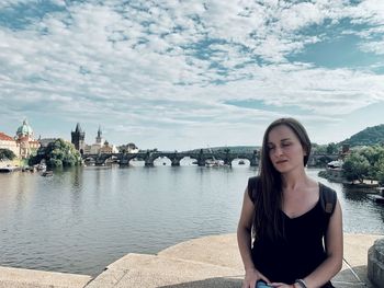 Portrait of young woman sitting on bridge over river against sky