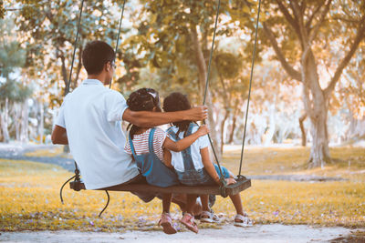 Rear view of father sitting with daughters on swing at park