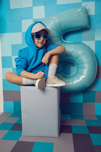 Boy child in blue clothes and a cap is sitting on a gray square celebrating his birthday. 