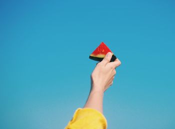 Cropped hand holding melon slice against clear blue sky