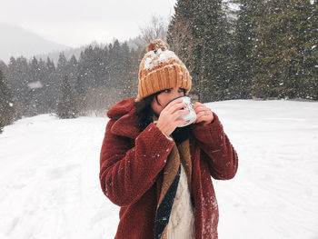 Happy young woman in winter clothes holding cup of hot tea outdoors, snowing, snow, nature.