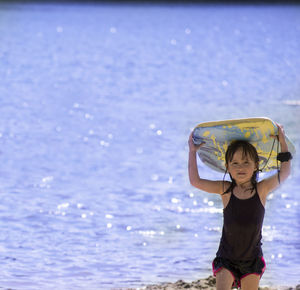 Portrait of girl carrying surfboard on head while standing at beach during sunny day