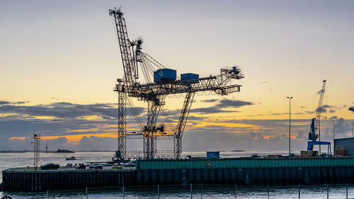 Cranes at commercial dock against sky during sunset