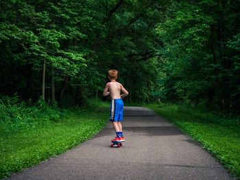 Rear view full length of shirtless boy skateboarding amidst trees on road