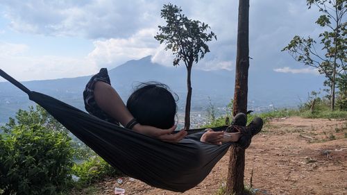 Woman sitting on land by trees against sky