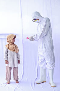 Full length of man wearing protective suit holding mask standing with cute girl against white background