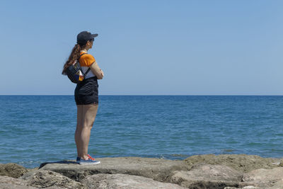 Side view of woman standing on rock while looking at sea against clear sky