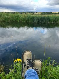 Low section of person standing on grass by lake against sky