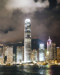 Illuminated city at waterfront against cloudy sky