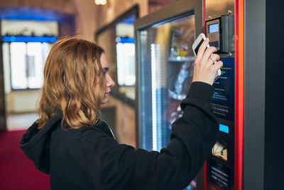 Woman paying for product at vending machine using contactless method of payment with mobile phone