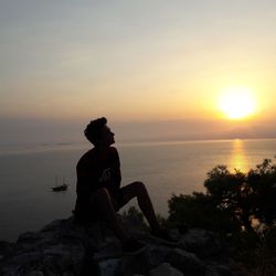 Man sitting on rock by sea against sky during sunset