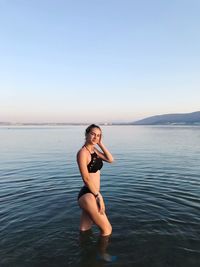 Portrait of young woman wearing bikini while standing in sea against clear sky at sunset