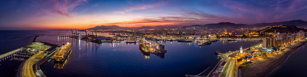 Panoramic view of illuminated city by sea at sunset