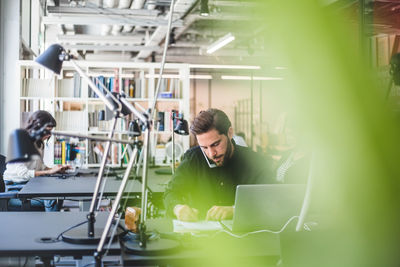 Businessman writing on paper while talking through mobile phone in creative office