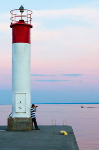 Full length of man by lighthouse photographing sea during sunset