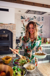 Female cook adding quinoa from frying pan in healthy salad with vegetables in light kitchen