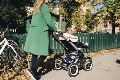 Mother pushing daughter in carriage while walking by fence in city