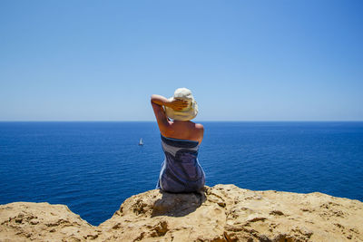 Rear view of woman sitting on rock by sea against sky