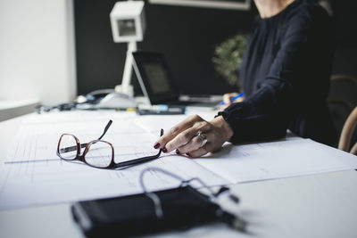 Midsection of female engineer holding eyeglasses on blueprint at desk