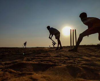 Silhouette of men playing cricket at beach against sky during sunset