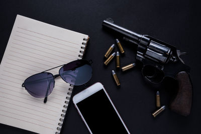 Close-up of sunglasses on spiral notebook by weapon and smart phone over black background