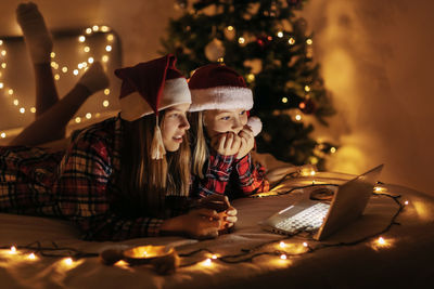 Two teenage girls in plaid shirts lying on the bed and using a laptop, a lighted christmas tree 