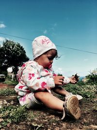 Side view of baby sitting on field against blue sky